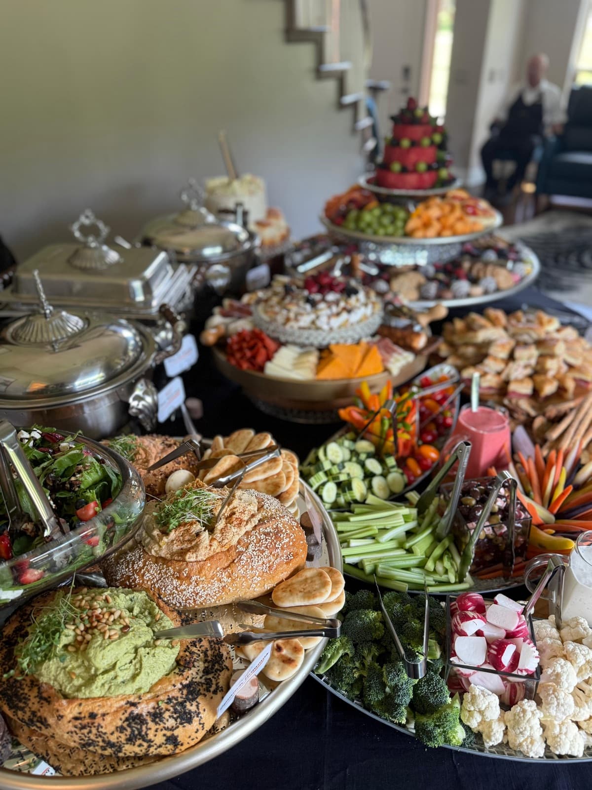 Generous catering spread with bread bowls of dip, fresh vegetables, and fruit