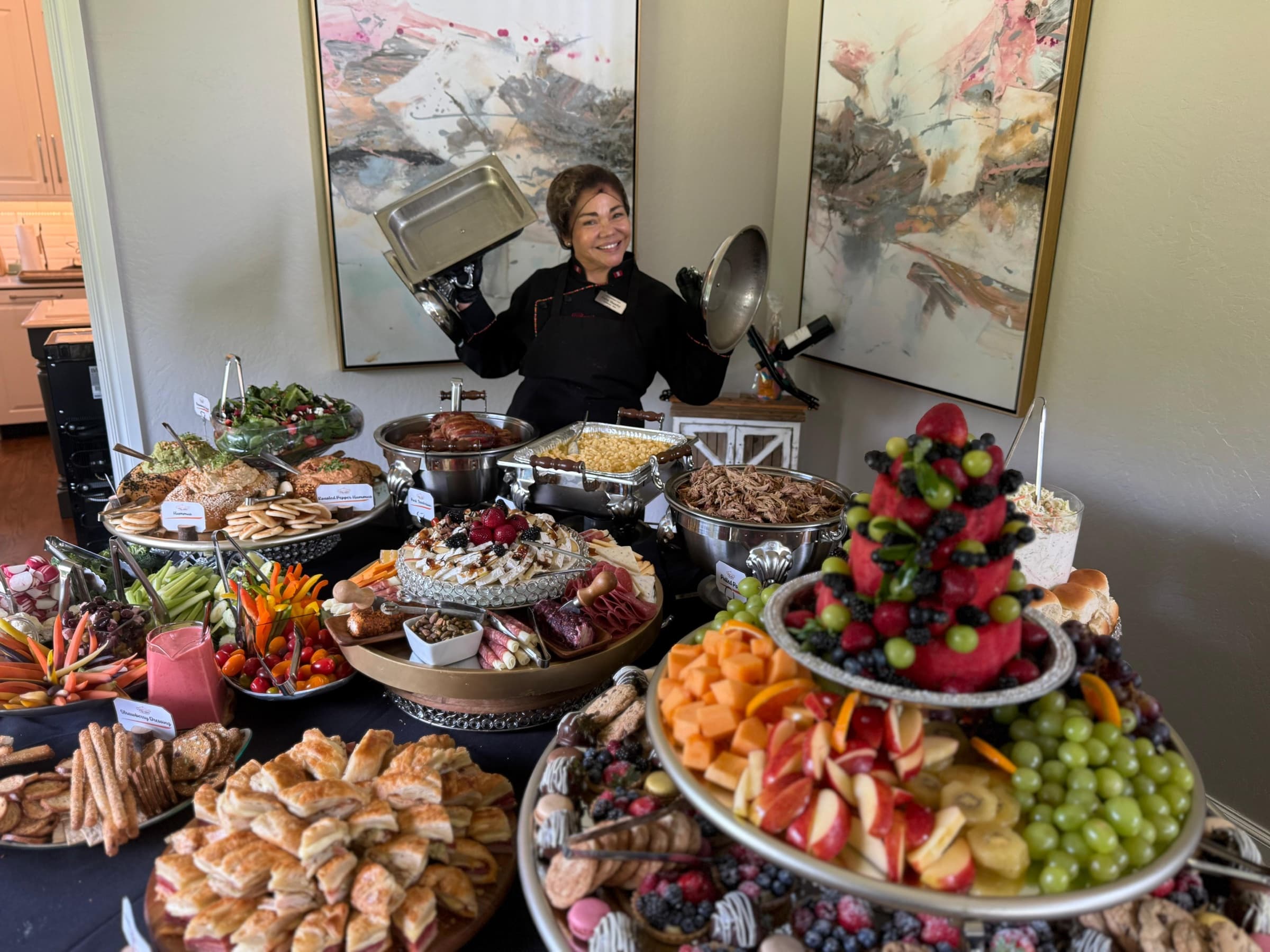 Patty standing behind an abundant catered spread of fresh fruit, charcuterie, salads, pastries, and warm dishes